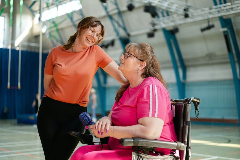 Two women laugh together in a community gym; one of them uses a wheelchair.